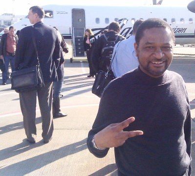 Get a quote today Man smiling and making a peace sign in front of an airplane with passengers in the background.